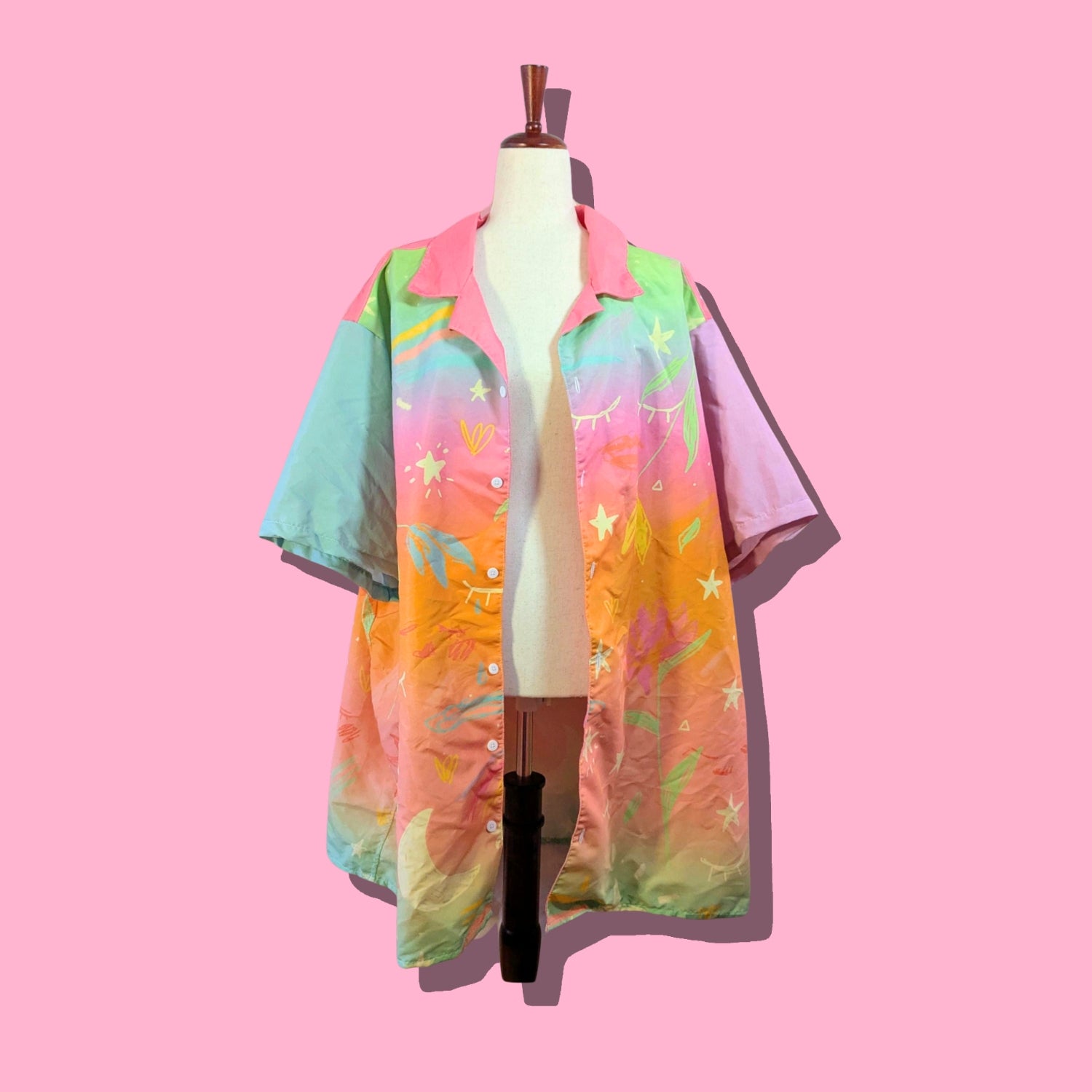 A short-sleeve button-up shirt with a vibrant pastel rainbow gradient and star and leaf patterns, displayed on a cream dress form mannequin against a solid pink background.