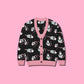 A black cardigan sweater featuring a playful pattern of cartoon clown opossums and pink trim, displayed flat against a solid pink background.