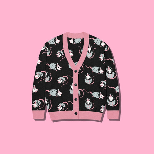 A black cardigan sweater featuring a playful pattern of cartoon clown opossums and pink trim, displayed flat against a solid pink background.